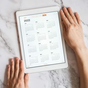 tablet computer with an open app of calendar for 2022 year in a womans hands on a gray marble background. concept business or to do list goals with technology using. top view, flat lay